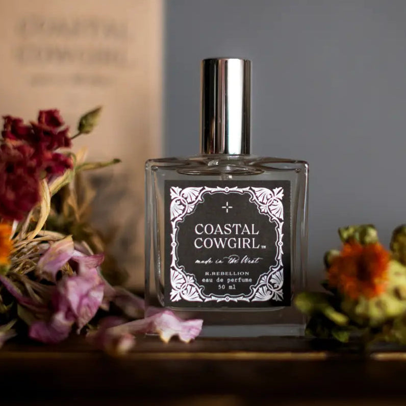 Perfume bottle labeled 'Coastal Cowgirl' surrounded by flowers on a wooden surface.
