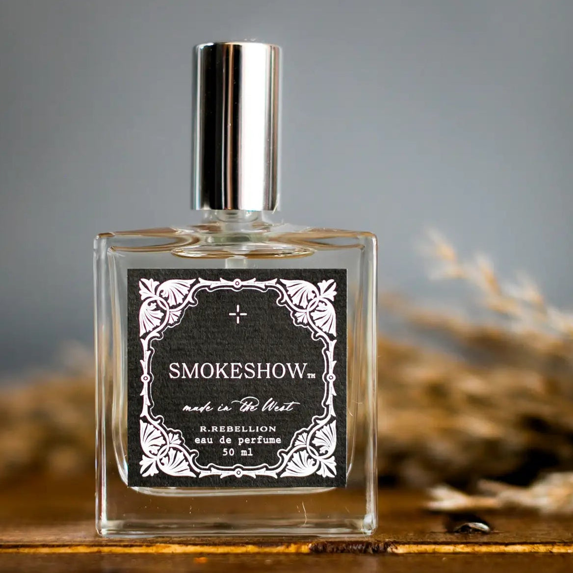 A glass bottle of 'Smoke Show' perfume with a black and white label, placed on a wooden surface with dried plants in the background.