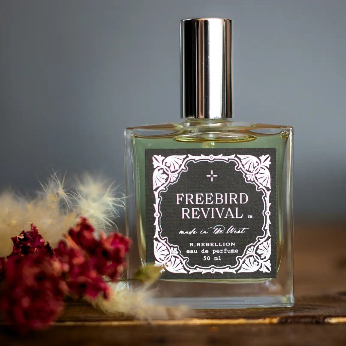 A clear glass bottle of Freebird Revival perfume with a silver cap, positioned on a wooden surface with a label that reads 'Freebird Revival' and '50 ml / 1.6 fl oz'.