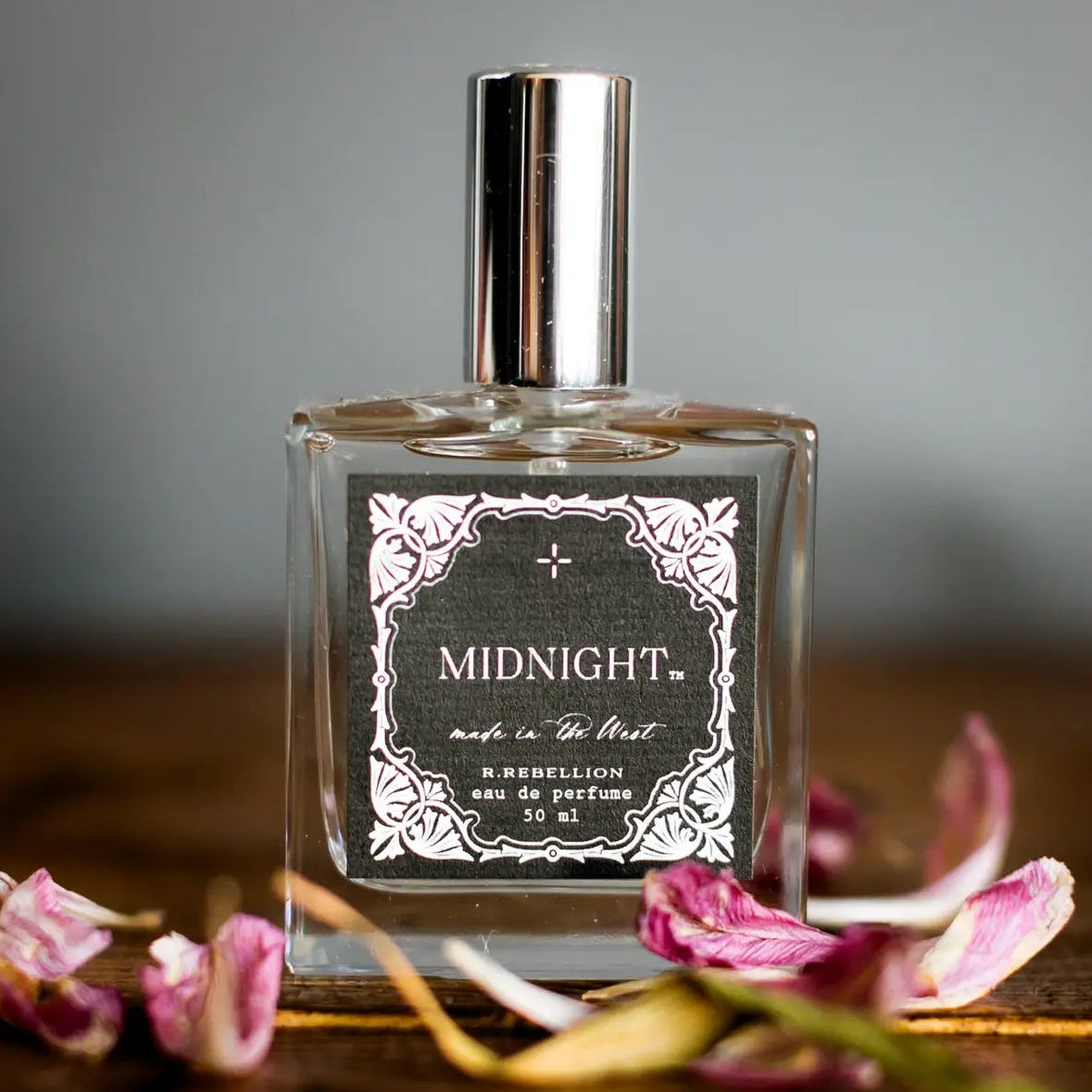 Midnight Perfume with a black and white western patterned label on a glass bottle with a tall silver cap, sitting on a wooden surface with dried flowers.