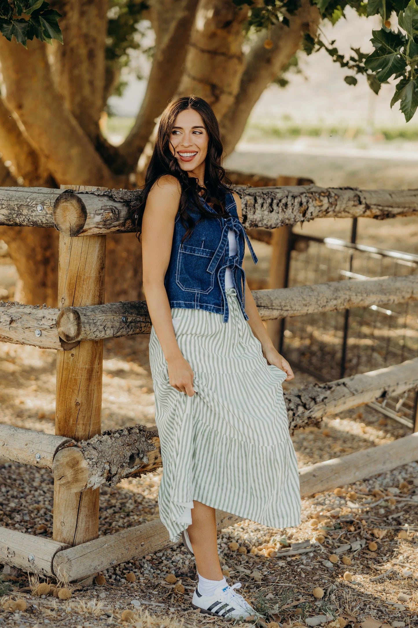 Woman in a denim top and striped skirt standing next to a wooden fence with trees in the background.