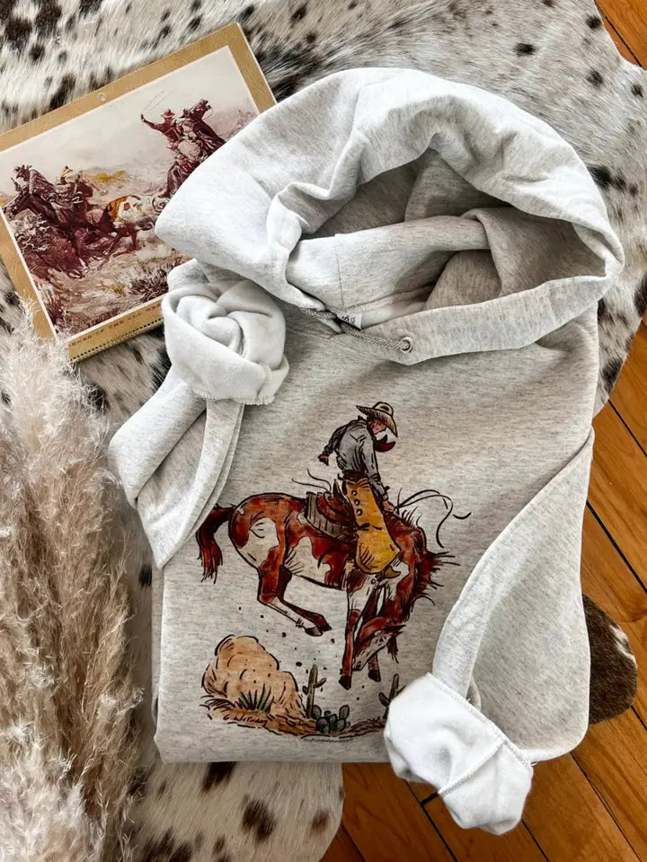 Light gray hoodie with a graphic of a cowboy on a horse, folded on a textured surface.